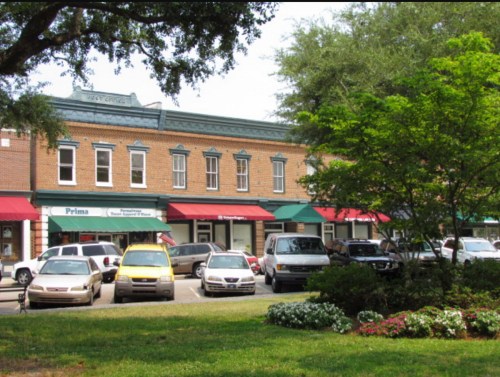 A row of brick storefronts with green awnings, parked cars in front, and lush greenery in the foreground.