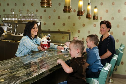 A woman serves a large ice cream sundae to two young boys at a counter, while a woman watches and smiles.