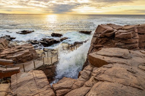 Waves crash against rocky shore at sunset, with wooden steps leading down to the water.