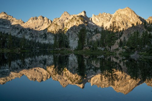 A serene mountain landscape reflecting in a calm lake, with towering peaks and lush trees under a clear blue sky.