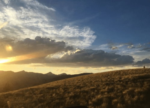 A scenic landscape at sunset, featuring rolling hills, clouds, and a warm golden glow in the sky.