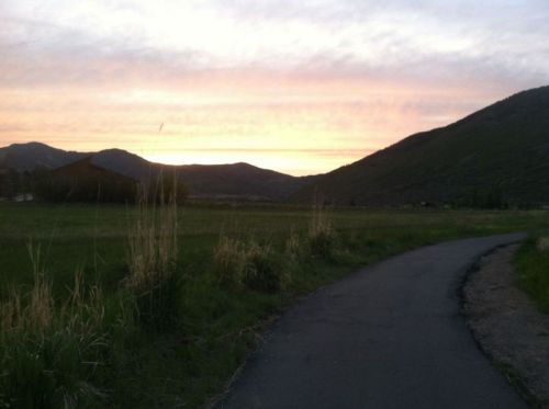 A winding path through a grassy field, with mountains and a colorful sunset in the background.