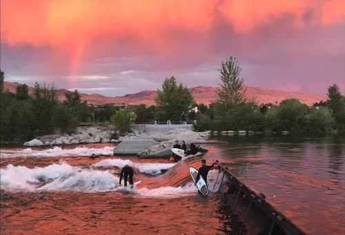 A vibrant sunset casts a pink hue over a river where surfers ride waves near rocky banks and lush trees.