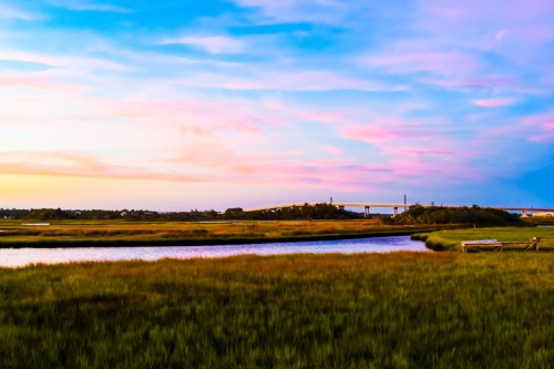 A serene landscape featuring a river, lush green grass, and a colorful sunset sky with a bridge in the background.