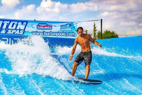 A shirtless surfer rides a wave on a blue surf simulator under a sunny sky.