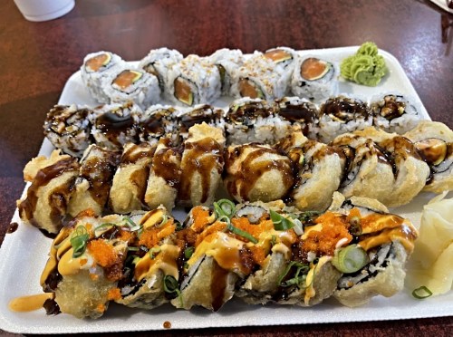 A platter of assorted sushi rolls, including tempura rolls, drizzled with sauces and garnished with wasabi and ginger.