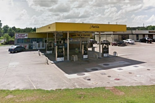 A yellow gas station with multiple fuel pumps, surrounded by shops and a grassy area under a cloudy sky.