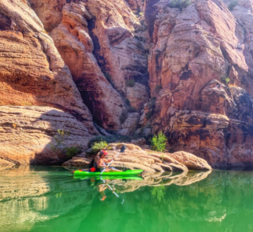 A person kayaking on calm green water surrounded by steep, rocky cliffs and lush greenery.