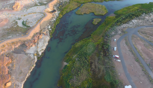 Aerial view of a winding river surrounded by green vegetation and rocky terrain, with vehicles parked nearby.
