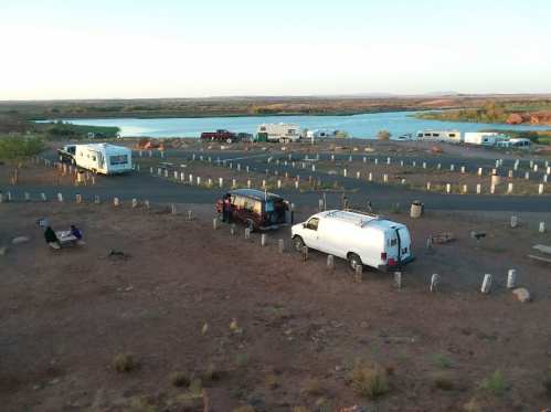 Aerial view of a campsite by a lake, featuring RVs and vehicles parked on a gravel lot surrounded by desert landscape.
