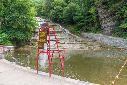 A swimming area with a sign, surrounded by lush greenery and a waterfall cascading down rocky terrain.