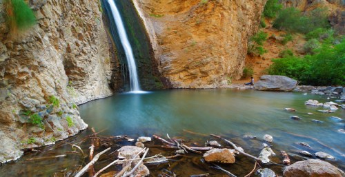 A serene waterfall cascades into a clear pool, surrounded by rocky cliffs and lush greenery.