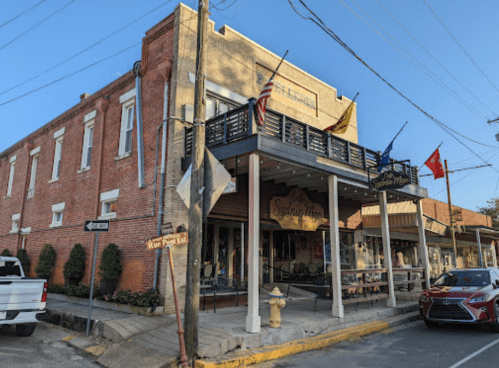 Historic brick building with a porch, flags, and shops in a small town setting. Street signs and parked cars visible.