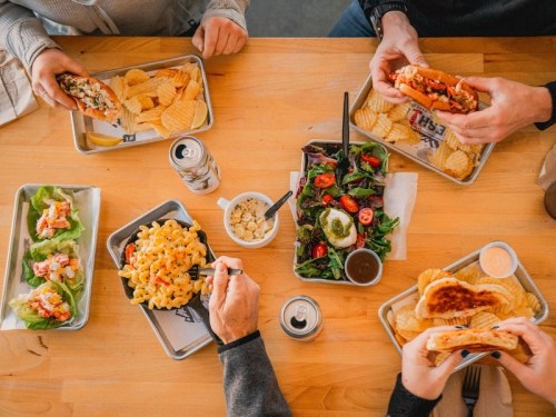 A table filled with various dishes: tacos, salads, pasta, chips, and drinks, with hands reaching for the food.