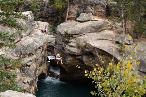Two people sit on rocky ledges by a river, surrounded by trees and boulders, with water flowing below.