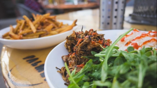 A plate of pulled meat with greens and rice, accompanied by a side of crispy fries on a wooden table.