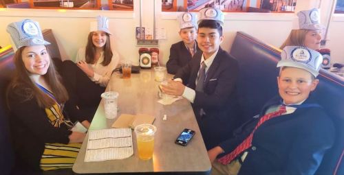 A group of six young people in formal attire wearing paper hats, sitting at a diner table with drinks and menus.