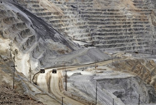 A large open-pit mine with steep, layered walls and trucks on a winding dirt road. Dust and rocky terrain are visible.