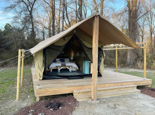 A glamping tent on a wooden platform, featuring a bed and rustic decor, surrounded by trees in a natural setting.