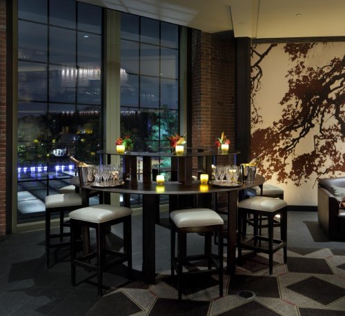 A modern bar area with a round table, stools, and decorative plants, illuminated by soft candlelight and large windows.