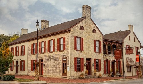 Historic stone building with red shutters, featuring a porch and signage, set in a quaint town.