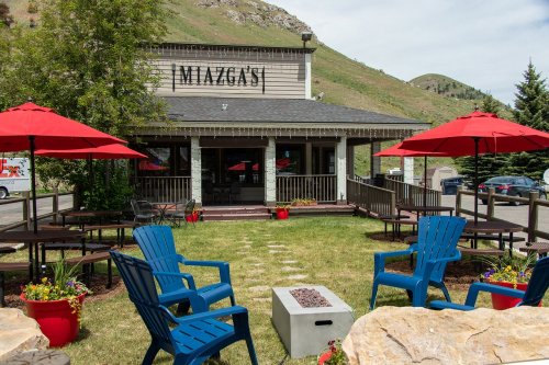 Outdoor seating area with blue chairs, red umbrellas, and a fire pit in front of a building named Miazga's, surrounded by greenery.