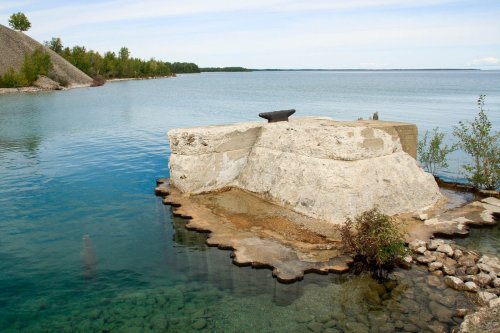 A rocky outcrop in calm water, surrounded by greenery and a clear blue sky.