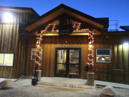 A wooden building entrance adorned with string lights, featuring double doors and a sign reading "The Den."