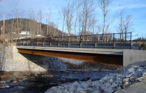 A concrete bridge spans a stream, surrounded by trees and a rural landscape under a clear blue sky.