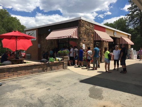 A busy ice cream shop with a long line of customers outside, shaded by red umbrellas on a sunny day.