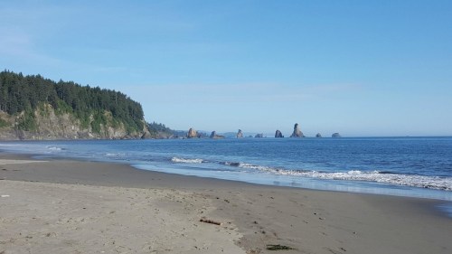 A serene beach scene with gentle waves, rocky formations in the distance, and lush green cliffs under a clear blue sky.