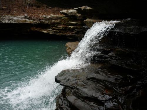 A small waterfall cascades over dark rocks into a serene turquoise pool surrounded by natural stone formations.