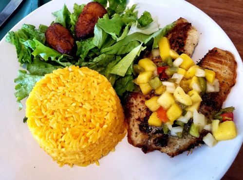 A plate featuring grilled fish topped with mango salsa, yellow rice, plantains, and a side of fresh greens.