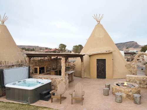 A cozy outdoor area featuring two teepee structures, a hot tub, and a stone fire pit surrounded by chairs.