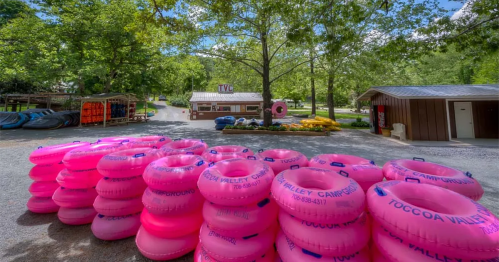 A stack of bright pink inner tubes in front of a rental shop surrounded by trees and colorful flowers.