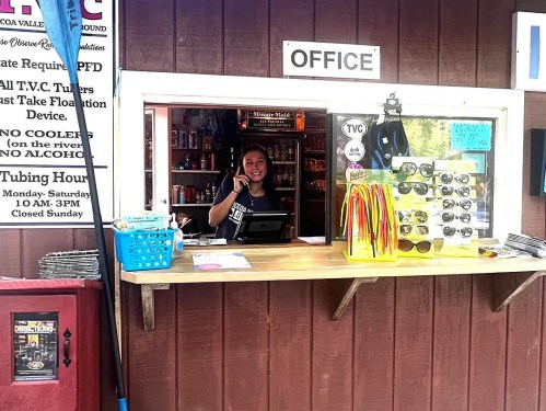 A smiling employee at a kiosk office, surrounded by sunglasses and colorful accessories, ready to assist customers.