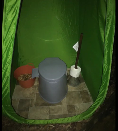 A green tent interior with a bucket, a gray container, a roll of paper towels, and a stick on a tiled floor.