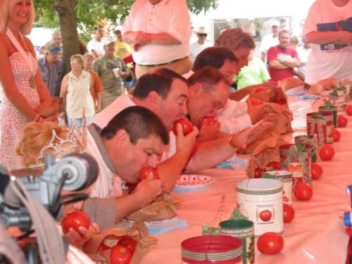 Participants compete in a tomato-eating contest, focused on their plates, with a crowd watching in the background.