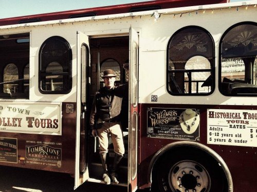 A man in period costume stands at the entrance of a vintage tour bus, promoting historical tours in Tombstone.