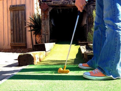 A person in jeans and orange sneakers prepares to putt on a mini-golf course with a cave entrance in the background.
