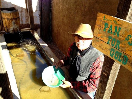 A man in a cowboy hat pans for gold in a wooden trough, with a sign reading "Learn to Pan for Gold" nearby.
