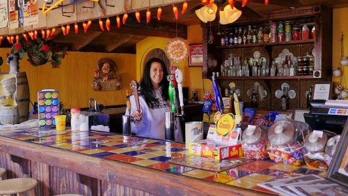 A smiling bartender stands behind a colorful bar filled with drinks and snacks, surrounded by festive decorations.