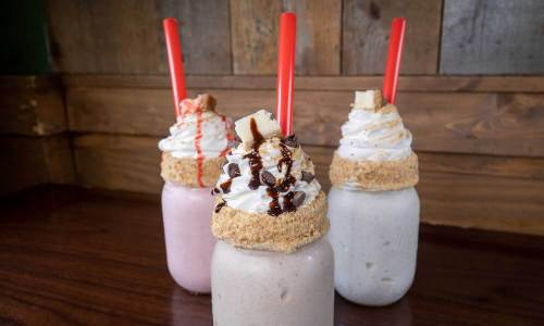 Three colorful milkshakes in jars topped with whipped cream, chocolate, and cookie crumbles, each with a red straw.