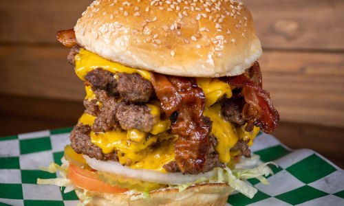 A towering cheeseburger with multiple beef patties, crispy bacon, melted cheese, lettuce, and tomato on a sesame seed bun.