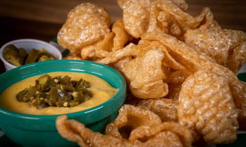A plate of crispy pork rinds served with a bowl of cheese dip and jalapeños on the side.