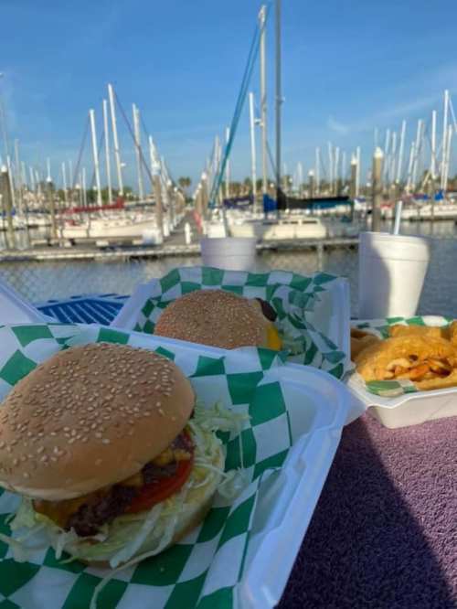 Two burgers on a table with a marina in the background, featuring boats and a clear blue sky.