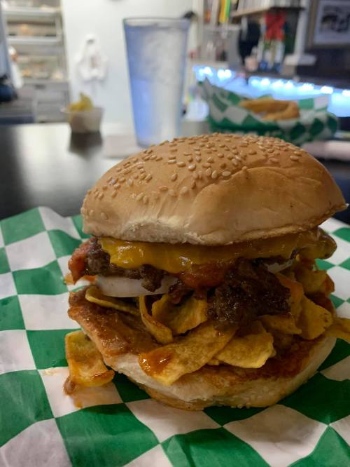 A close-up of a burger with cheese, toppings, and Fritos on a sesame seed bun, served on green and white checkered paper.