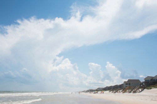 A serene beach scene with soft waves, sandy shore, and fluffy clouds under a bright blue sky. Beach houses line the coast.