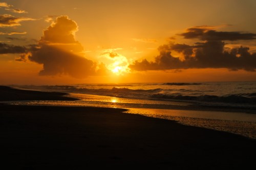 A serene beach at sunset, with golden hues reflecting on the water and clouds scattered in the sky.