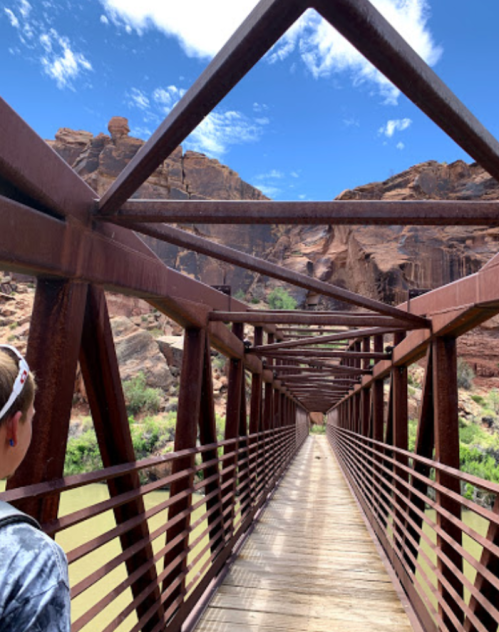 A person stands on a metal bridge, surrounded by rocky cliffs and a blue sky with scattered clouds.